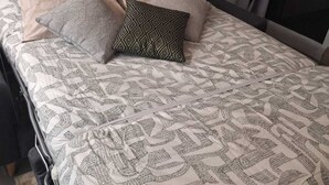 A bed with patterned bedding and pillows, a bedside table, and a window with blinds. - La parenthese suspendu (Font-Romeu-Odeillo-Via)