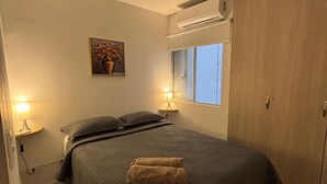 Un dormitorio con cama, lámparas de noche, un cuadro, un equipo de aire acondicionado y una ventana.