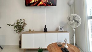 Una sala de estar moderna con un televisor de pantalla plana, un mueble blanco, una mesa redonda de madera, una pequeña planta en maceta y un ventilador de pie.
