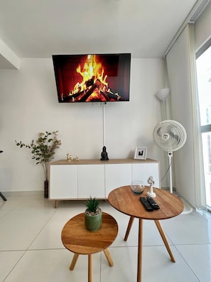 Una sala de estar moderna con un televisor de pantalla plana, un mueble blanco, una mesa redonda de madera, una pequeña planta en maceta y un ventilador de pie.
