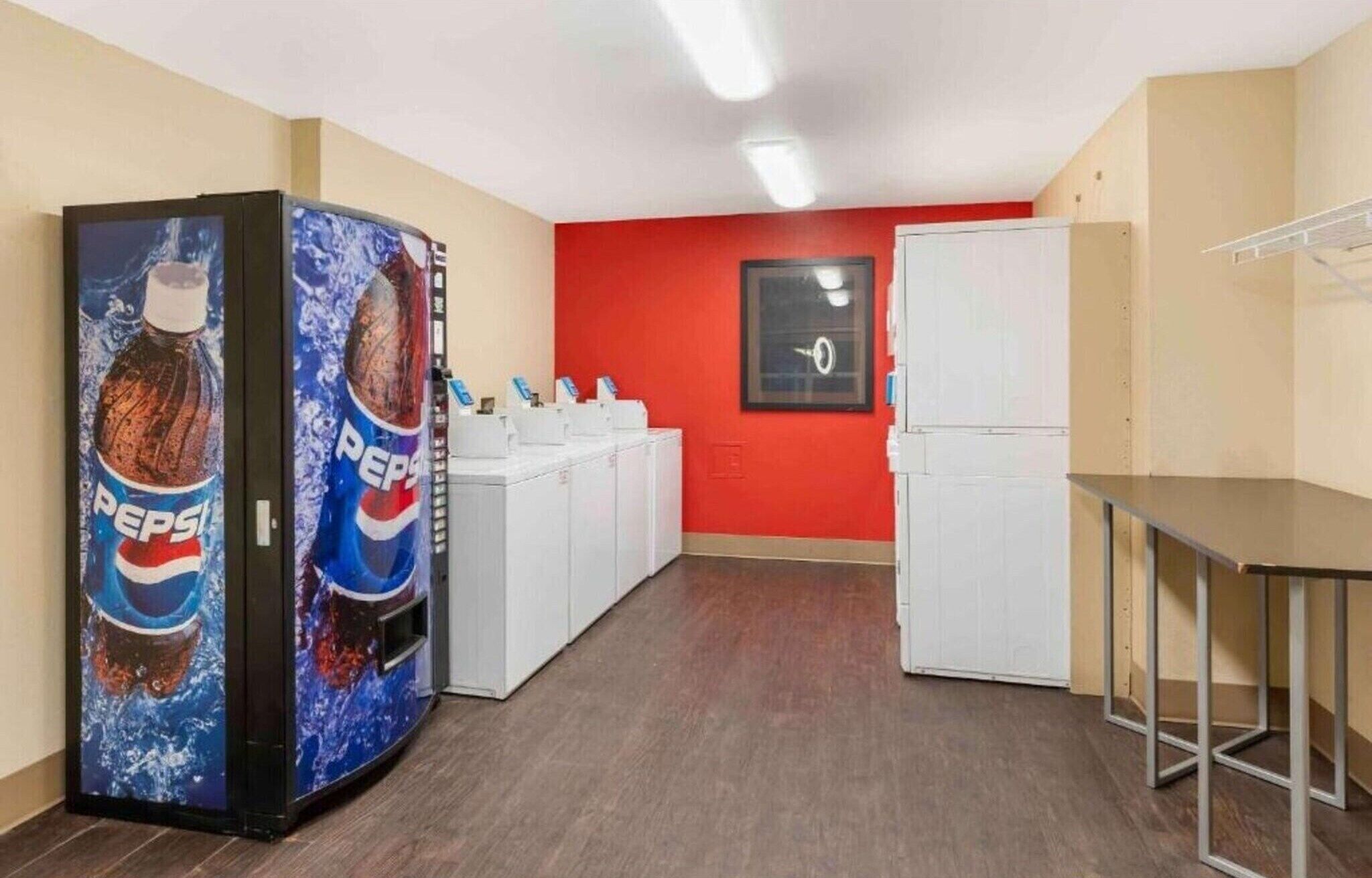 Ett rum med en röd vägg, ett vitt kylskåp, en vit tvättmaskin och två automater som säljer Pepsi.