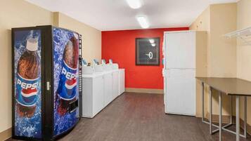 Ett rum med en röd vägg, ett vitt kylskåp, en vit tvättmaskin och två automater som säljer Pepsi.