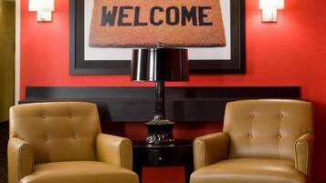 Un hall d’hôtel avec un mur rouge, un paillasson, deux fauteuils en cuir et une table d’appoint avec une lampe.