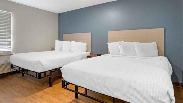 Dos camas individuales con cabeceros de madera y ropa de cama blanca en una habitación con una pared azul de acento y una ventana con persianas.