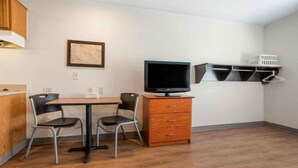 A small room with a wooden table, two chairs, a TV on a wooden cabinet, a painting on the wall, and a shelf with baskets. - ADA Full Bed,Tub,Nsmk|Kitchen|Studio 6 Fort Myers (Fort Myers FL)