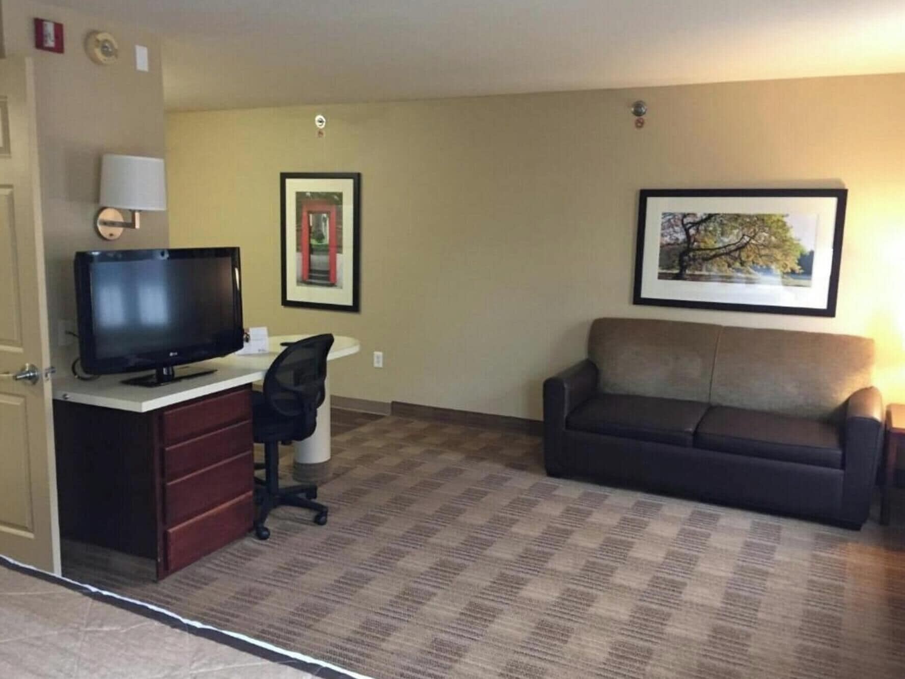 Habitación de hotel con televisión sobre un escritorio, un sofá, un cuadro en la pared y suelo alfombrado.