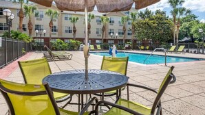 Zona junto a la piscina con mesa y sillas, piscina y un edificio con varias ventanas, con palmeras al fondo.