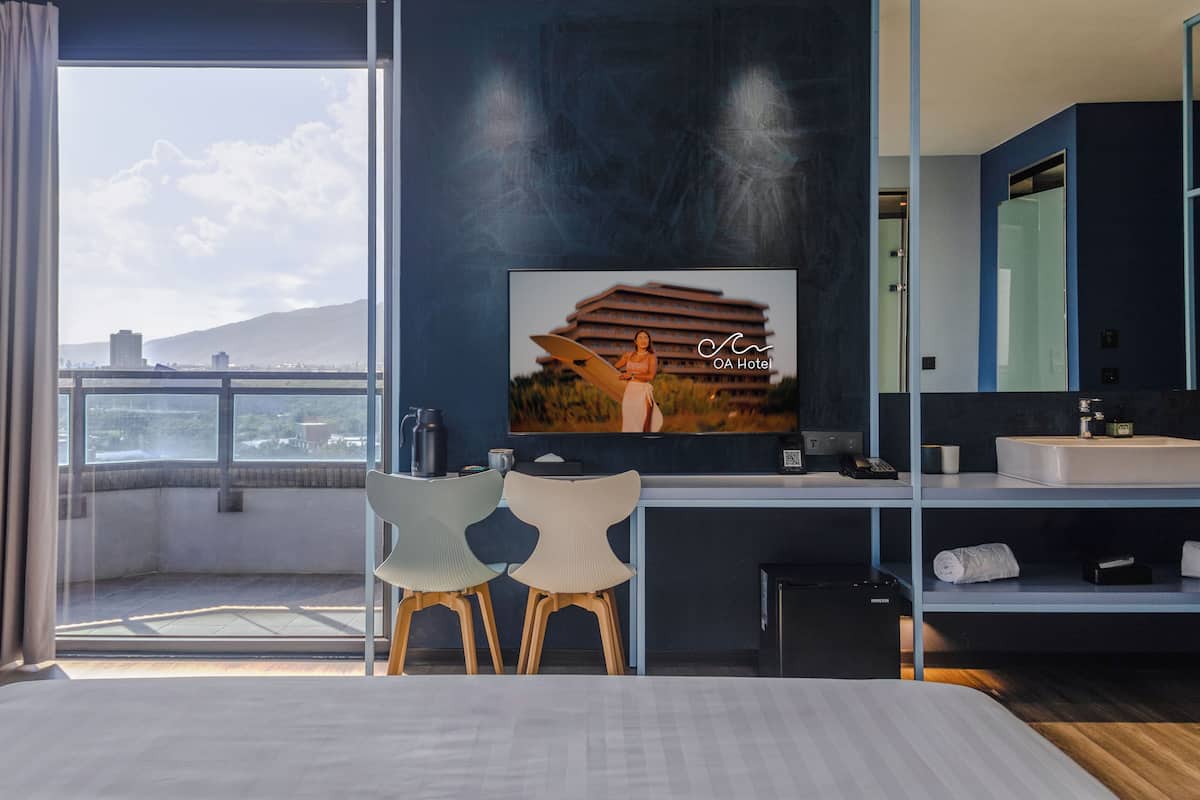 A hotel room with a large window, a bed, a desk with a chair, a bathroom with a mirror, and a painting on the wall.