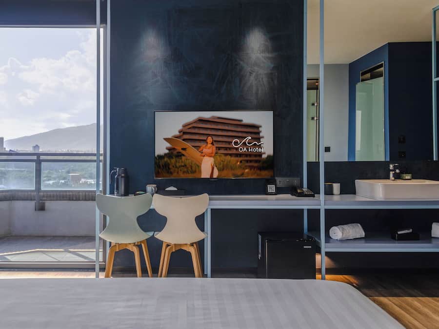 A hotel room with a large window, a bed, a desk with a chair, a bathroom with a mirror, and a painting on the wall.