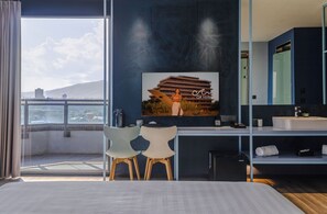 Ein Hotelzimmer mit großem Fenster, einem Bett, einem Schreibtisch mit Stuhl, einem Badezimmer mit Spiegel und einem Bild an der Wand.
