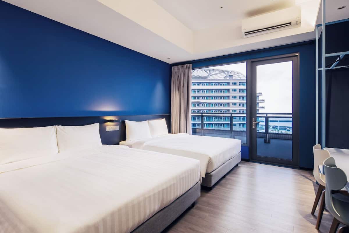 A hotel room with two beds, a large window with a view of a building, and a balcony.