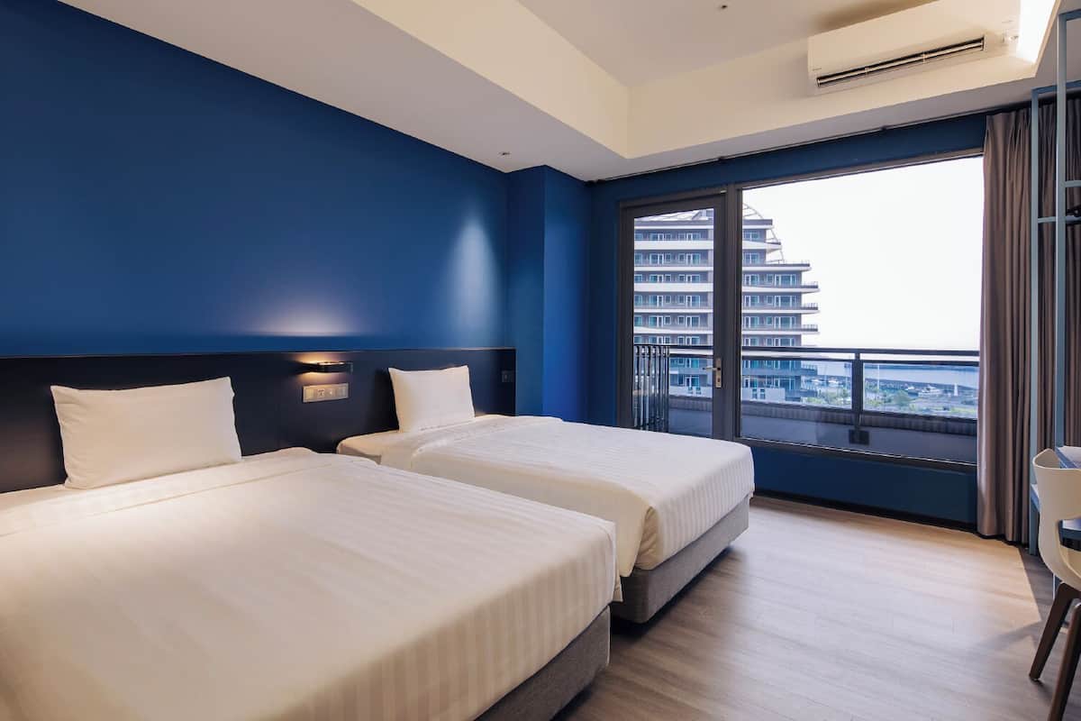 A hotel room with two beds, a large window with a view of buildings, and an air conditioning unit.