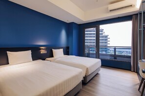 Ein Hotelzimmer mit zwei Betten, einem großen Fenster mit Blick auf Gebäude und einer Klimaanlage.