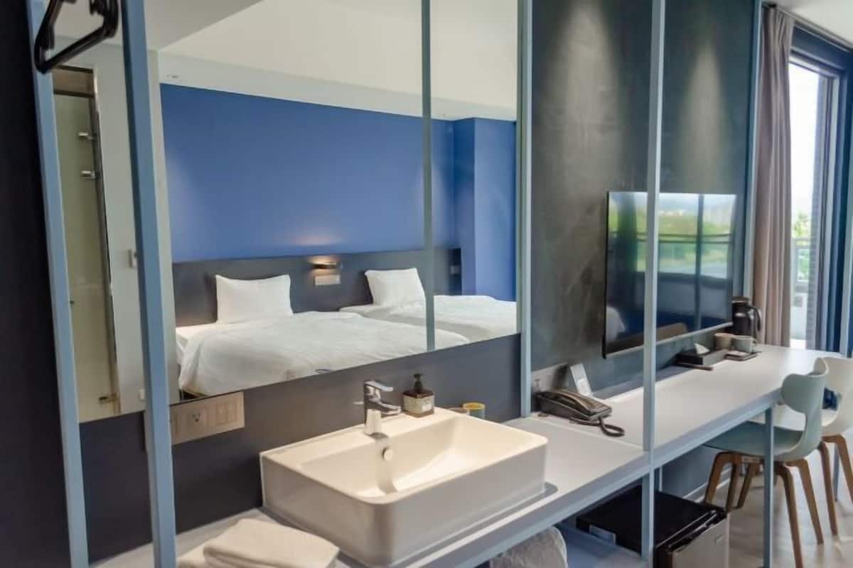 A modern hotel room with a large mirror, a white sink, a bed with white linens, and a desk with a chair.