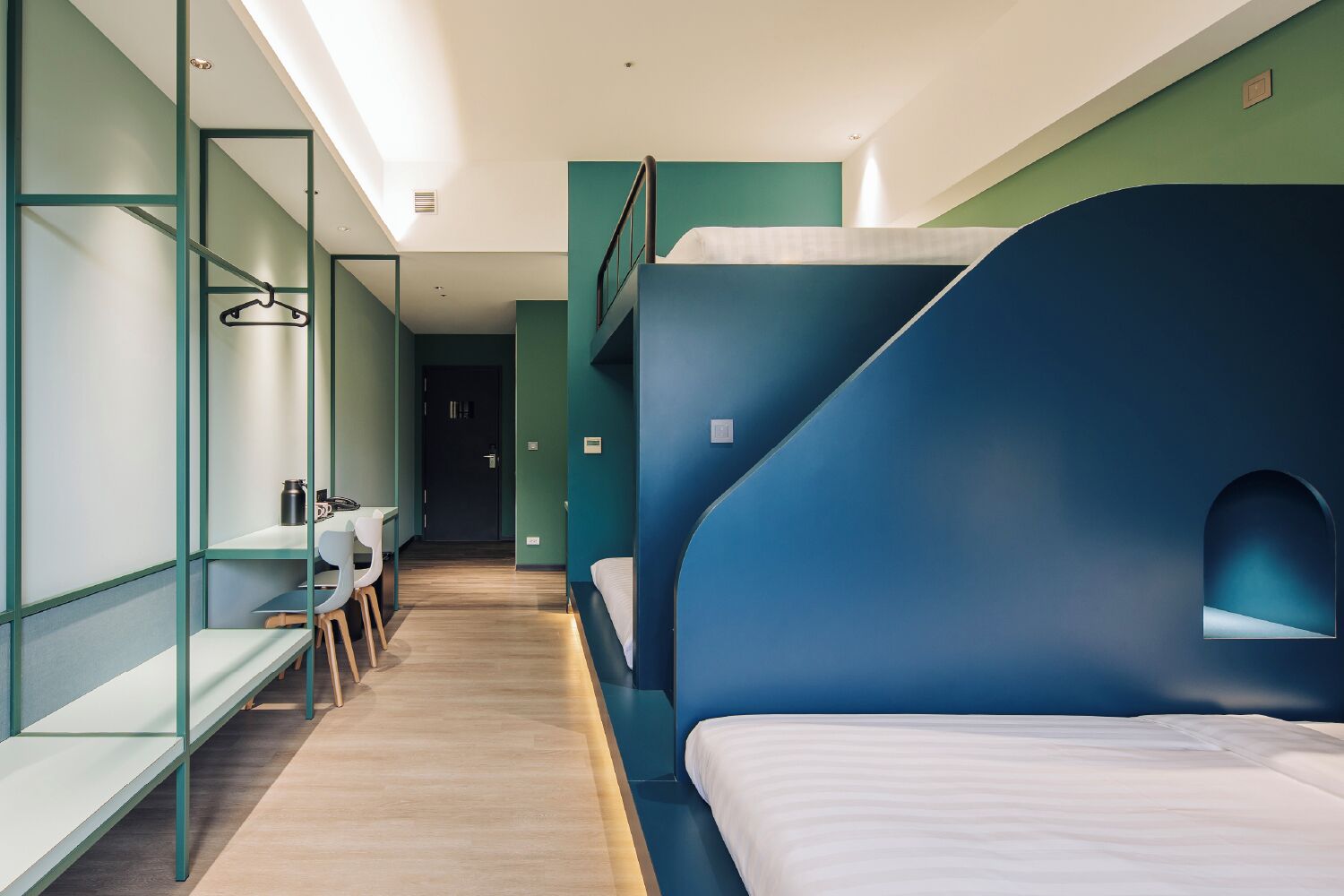 A modern hotel room with a bunk bed, a desk with a chair, a hanging coat rack, and a small window.