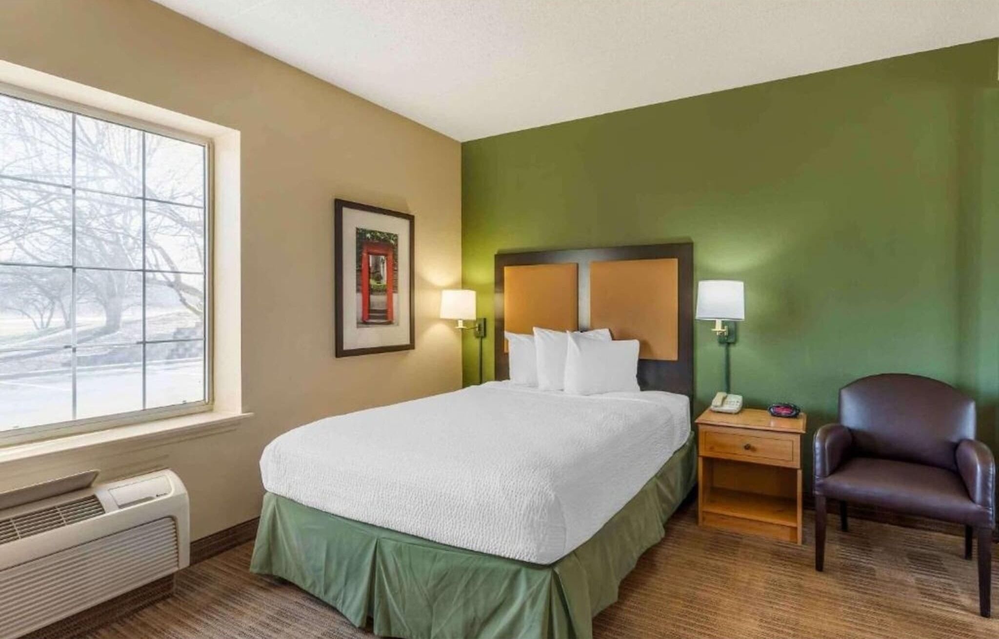 Une chambre d’hôtel avec un lit, une chaise, une table de chevet avec une lampe, un tableau encadré au mur et une fenêtre donnant sur des arbres.