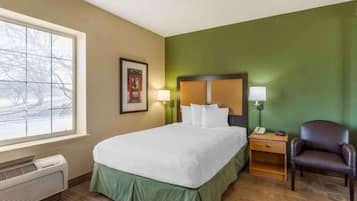 Une chambre d’hôtel avec un lit, une chaise, une table de chevet avec une lampe, un tableau encadré au mur et une fenêtre donnant sur des arbres.