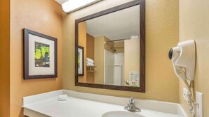 A hotel bathroom with a white sink, a large mirror, a framed picture on the wall, and a hairdryer on the wall. - Studio 6 Vernon Hills|ADA Queen bed,RI Shower,Nsmk|Kitchen (Chicago)
