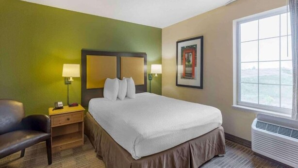 A hotel room with a bed, two bedside lamps, a chair, a nightstand, a framed picture on the wall, and a window with a view. - Studio 6 Vernon Hills|ADA Queen bed,RI Shower,Nsmk|Kitchen (Chicago)