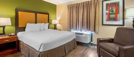 Habitació d'hotel amb un llit, taules de nit, una cadira, una finestra amb cortines, aire condicionat i un quadre amb marc a la paret.