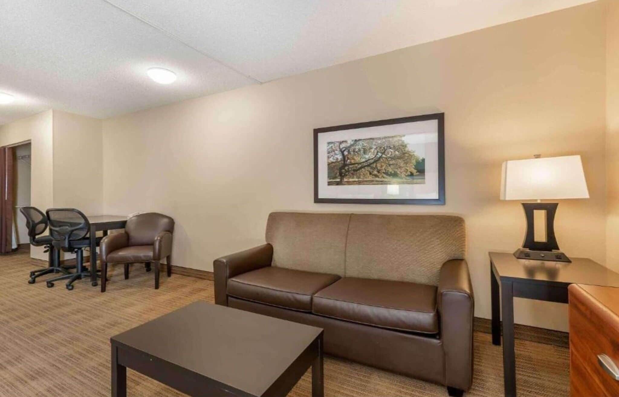 A hotel room with a sofa, a small table, a desk with a chair, a lamp, and a framed picture on the wall.