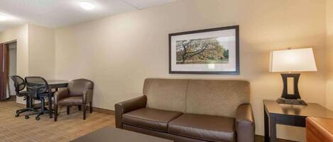 A hotel room with a sofa, a small table, a desk with a chair, a lamp, and a framed picture on the wall.