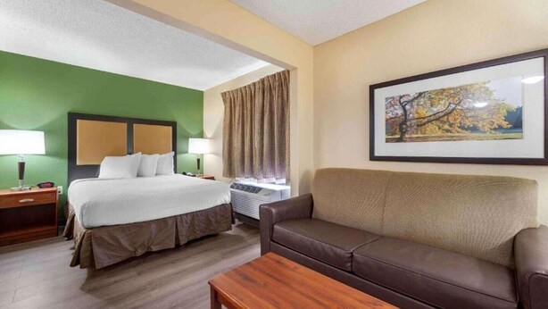 Une chambre d’hôtel avec un lit, un canapé, une table, une fenêtre avec des rideaux et un tableau encadré au mur.
