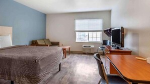 A hotel room with a bed, a desk with a computer, a chair, a window with blinds, and an air conditioning unit.