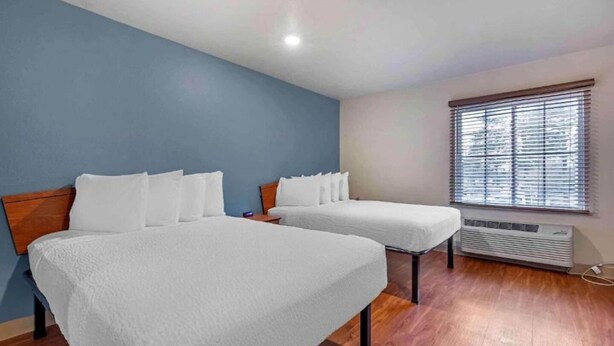 A room with two beds, a window with blinds, and a wall-mounted air conditioner.