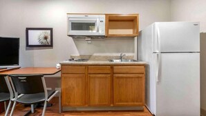 A compact kitchen with a microwave, sink, and refrigerator, wooden cabinets, and a small dining area with a chair.