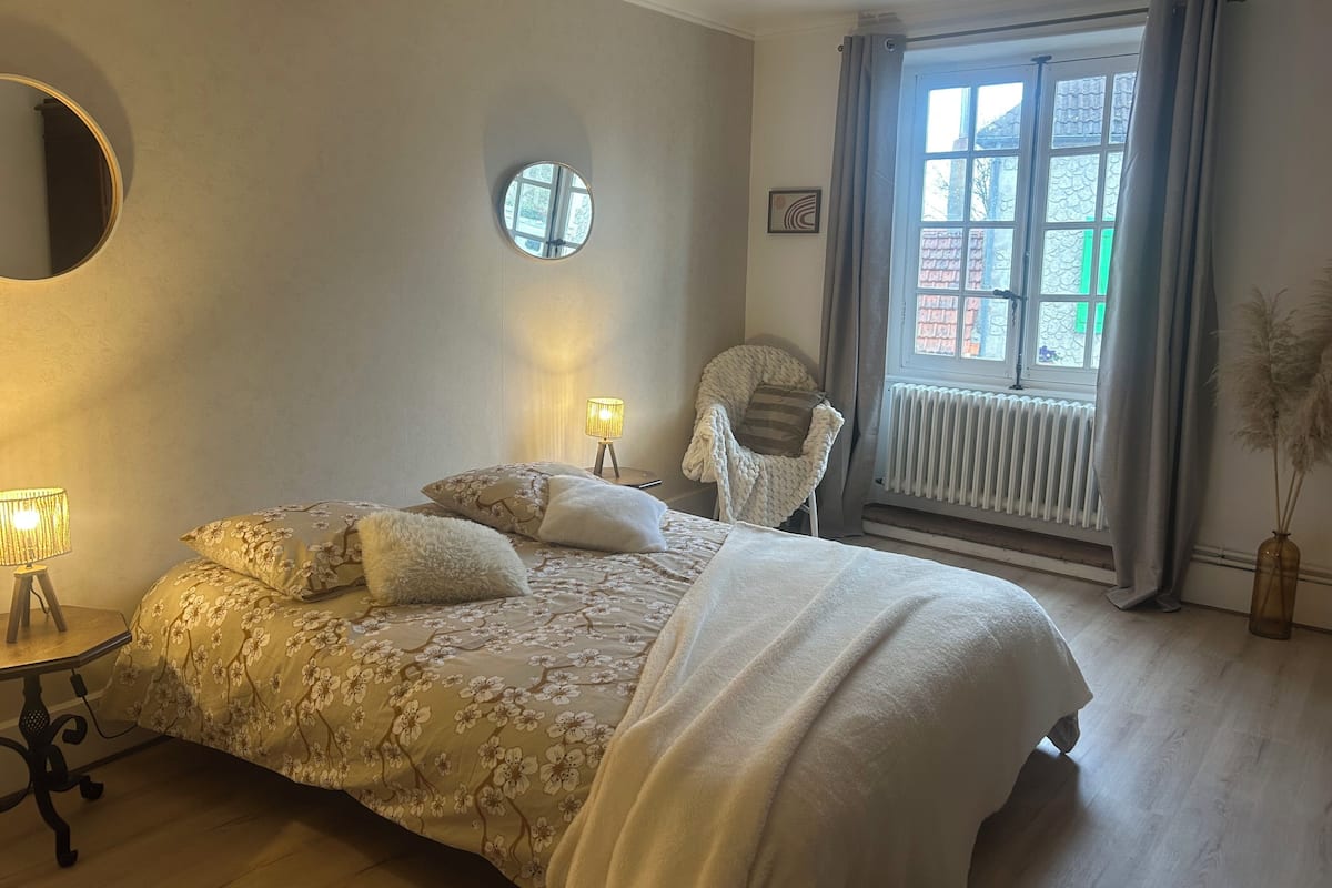 Une chambre à coucher avec un lit, un miroir rond, une chaise, une table avec une lampe et une fenêtre avec des rideaux.