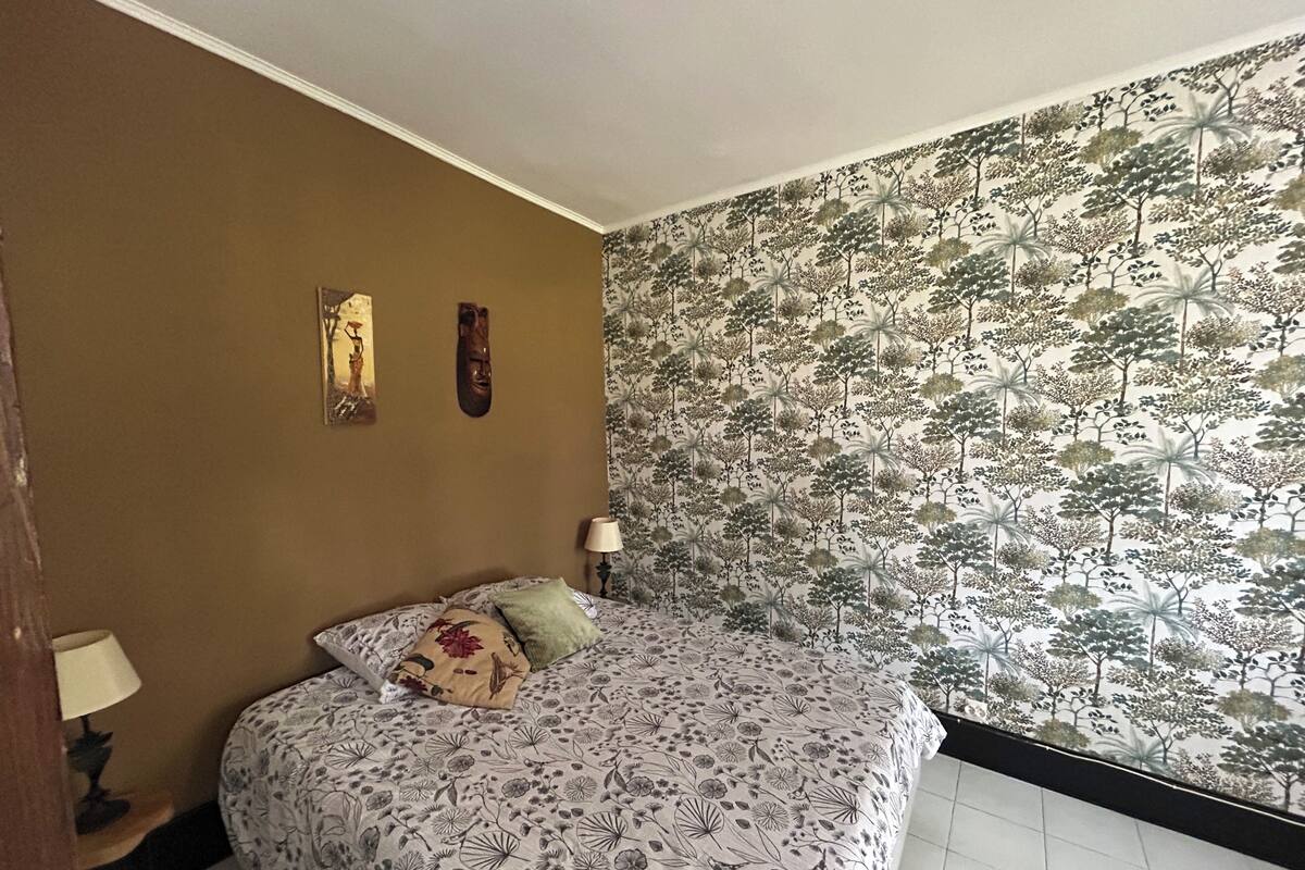 Une chambre à coucher comprenant un lit, une table de chevet, une lampe et un mur recouvert d’un papier peint à motifs floraux.