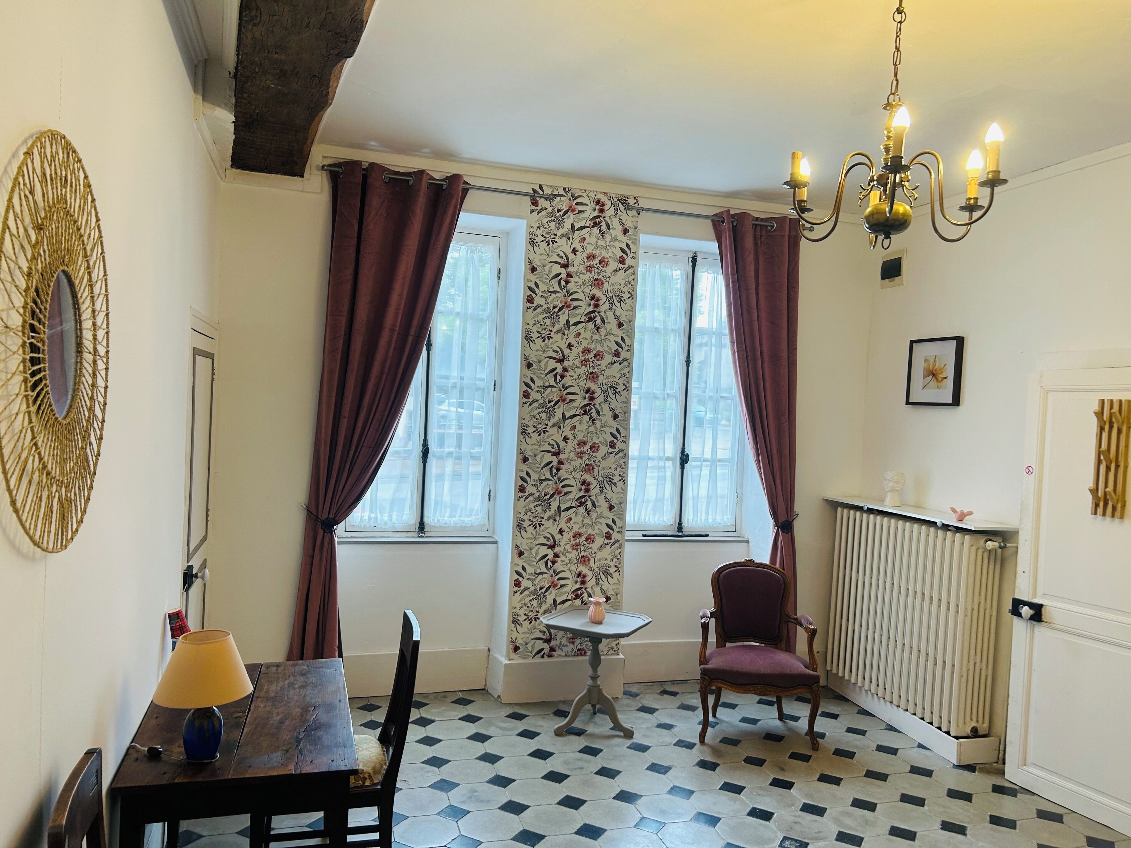 Een kamer met een houten tafel, een stoel, een bijzettafeltje met een vaas, een stoel met een kussen, een radiator, een raam met gordijnen en een kroonluchter.