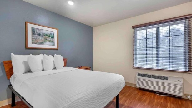 A bedroom with a bed, a framed picture on the wall, a window with blinds, and a wall-mounted air conditioner. - 1 Full Bed | Non-Smoking, Kitchen (Shreveport LA)