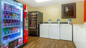 Um quarto com uma máquina de venda automática de Coca-Cola, uma máquina de autoatendimento para lanches e bebidas, e uma fileira de máquinas de lavar roupa.