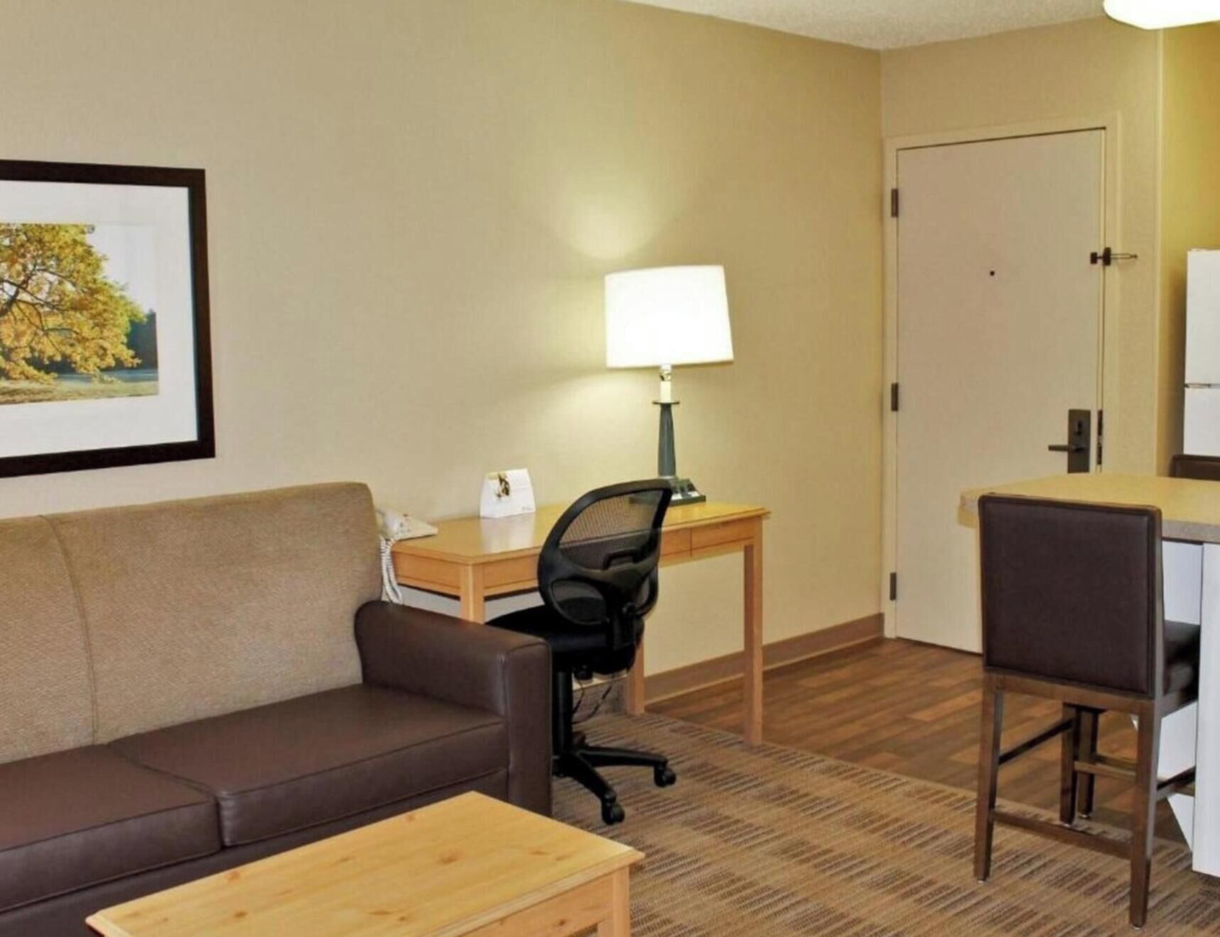 Una habitación de hotel con un sofá de cuero marrón, un escritorio de madera con una lámpara, una computadora y una silla.