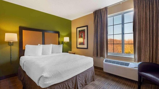 A hotel room with a bed, a nightstand, a lamp, a framed picture, a window with curtains, and a chair. - 1 King Bed | Non-Smoking, Kitchen (Minneapolis MN)