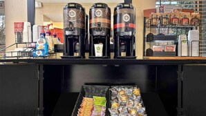 Una estación de café con tres dispensadores de café, un letrero de salida de incendios y una exhibición de alimentos envasados.