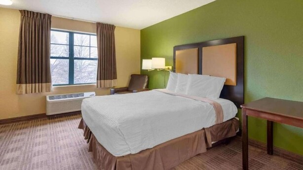 Habitación de hotel con cama, una silla, escritorio, ventana con cortinas y una lámpara fijada a la pared.