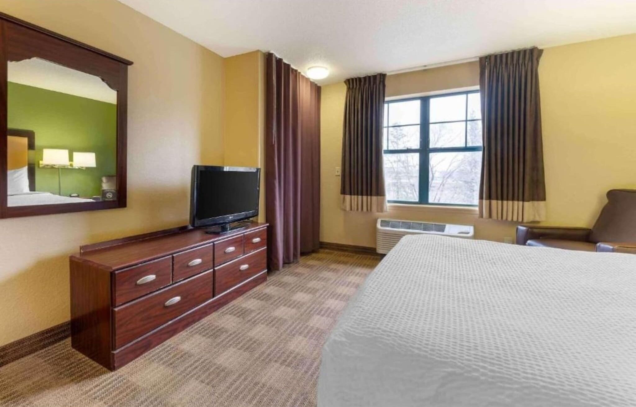A hotel room with a bed, a TV on a wooden dresser, a window with curtains, and a mirror on the wall.