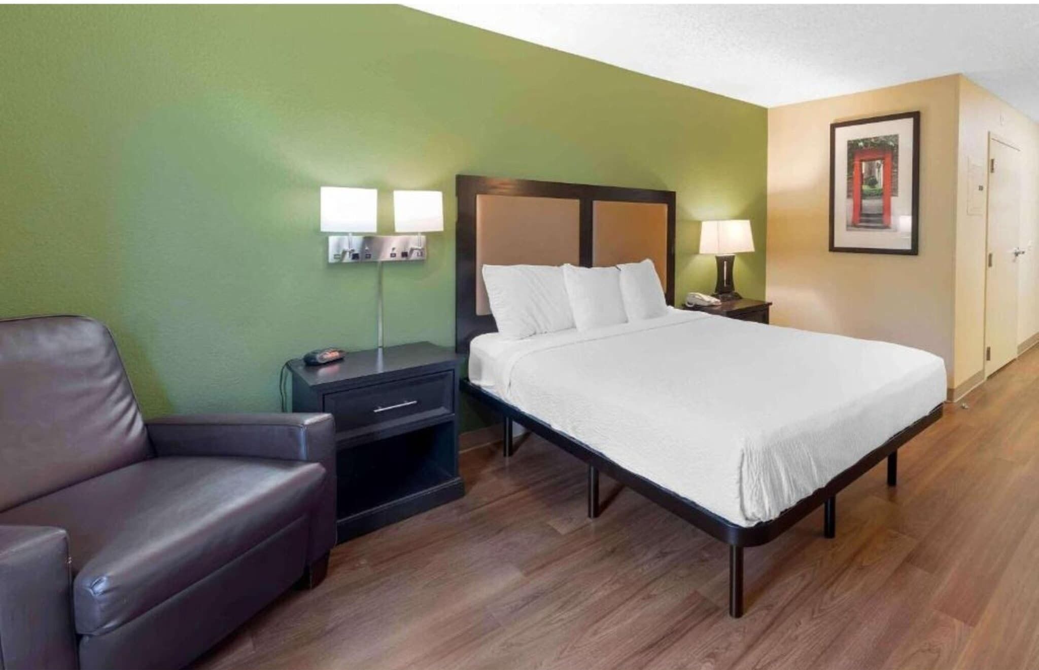 Een hotelkamer met een bed, een stoel, een nachtkastje, een lamp en een lijst met een afbeelding aan de muur.