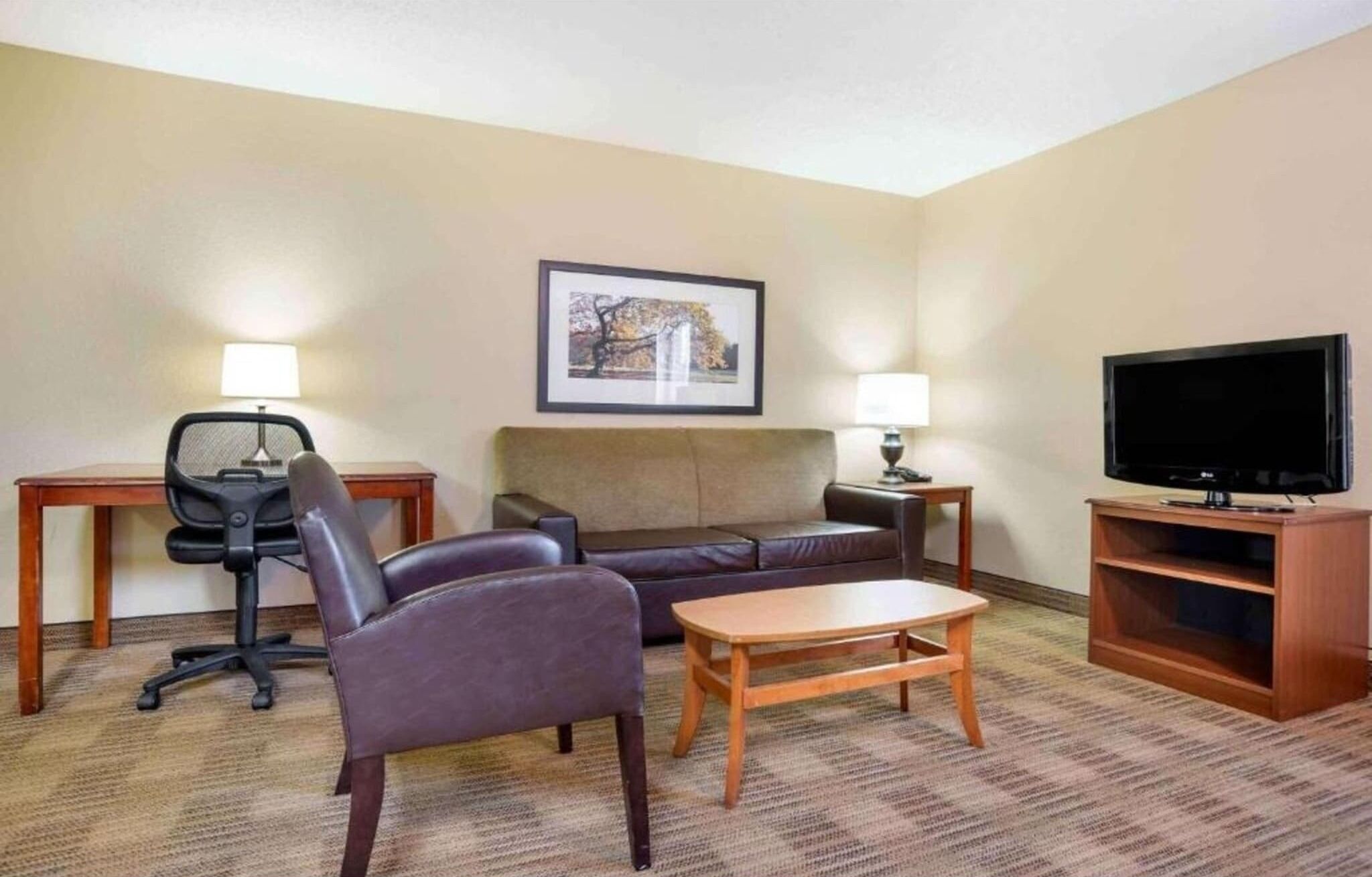 Une chambre d’hôtel avec un canapé, un fauteuil, une petite table, un téléviseur, une lampe et un tableau encadré au mur.