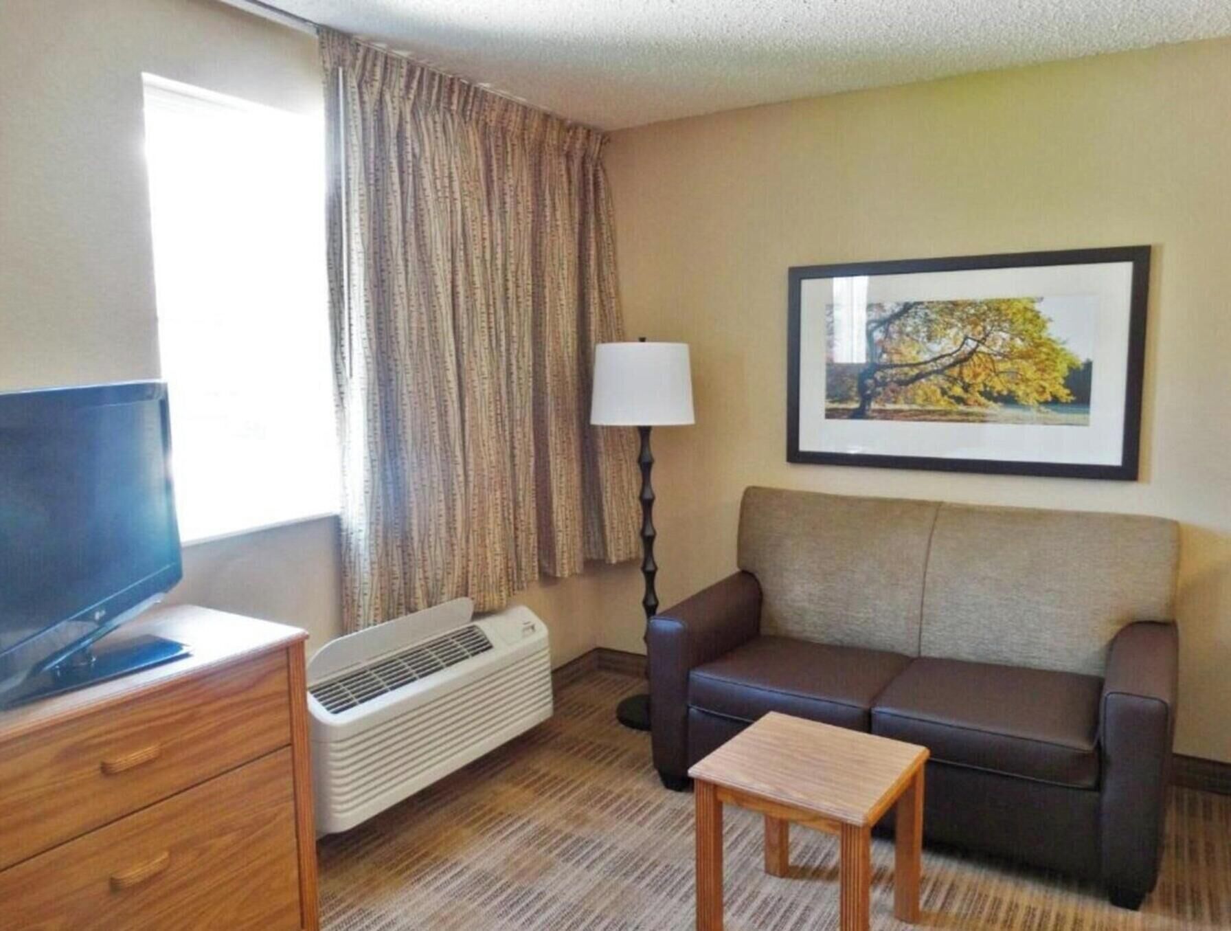 Een hotelkamer met een televisie op een houten dressoir, een sofa, een klein houten tafeltje, een lamp en een lijstje met een foto aan de muur.