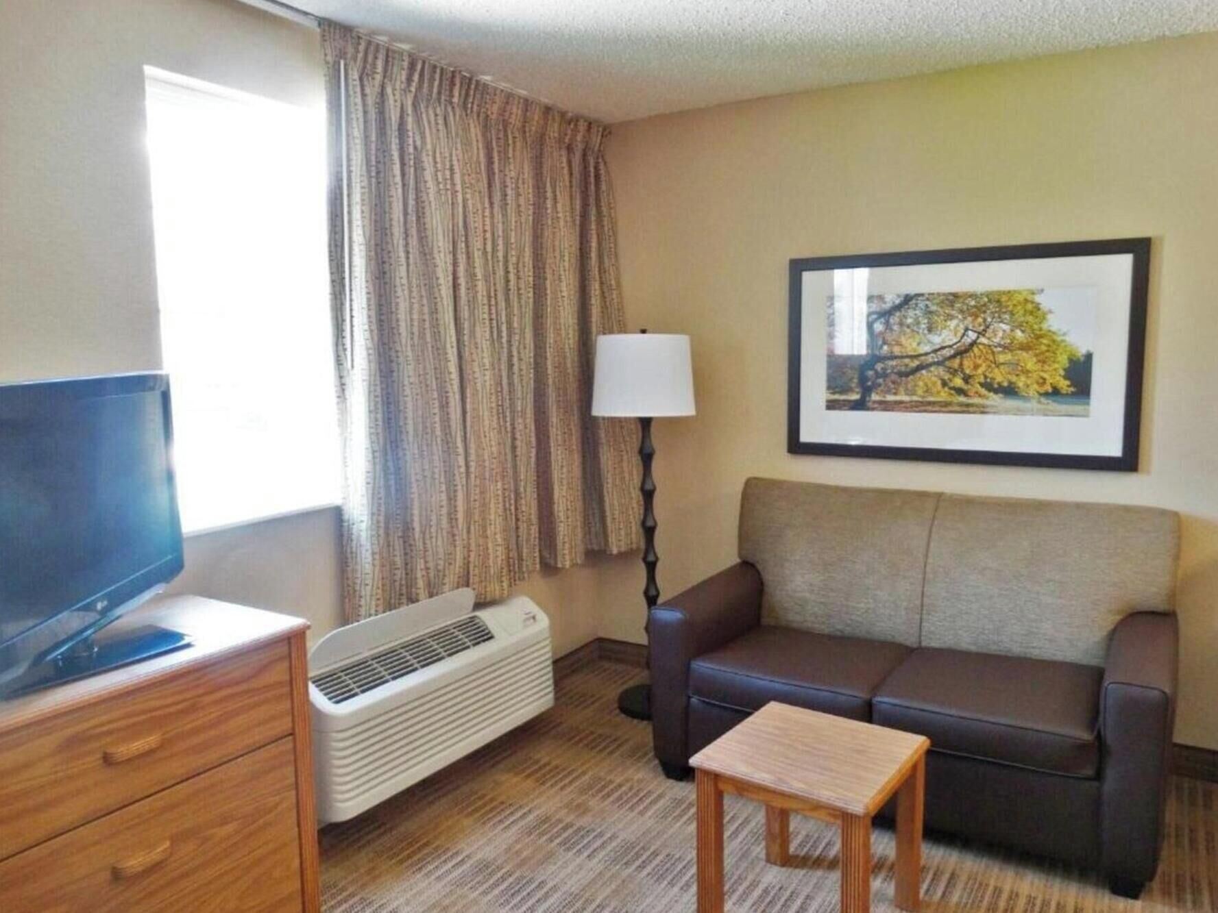 Ein Hotelzimmer mit Fernseher auf einer Holzkommode, einem Sofa, einem kleinen Holztisch, einer Lampe und einem gerahmten Bild an der Wand.