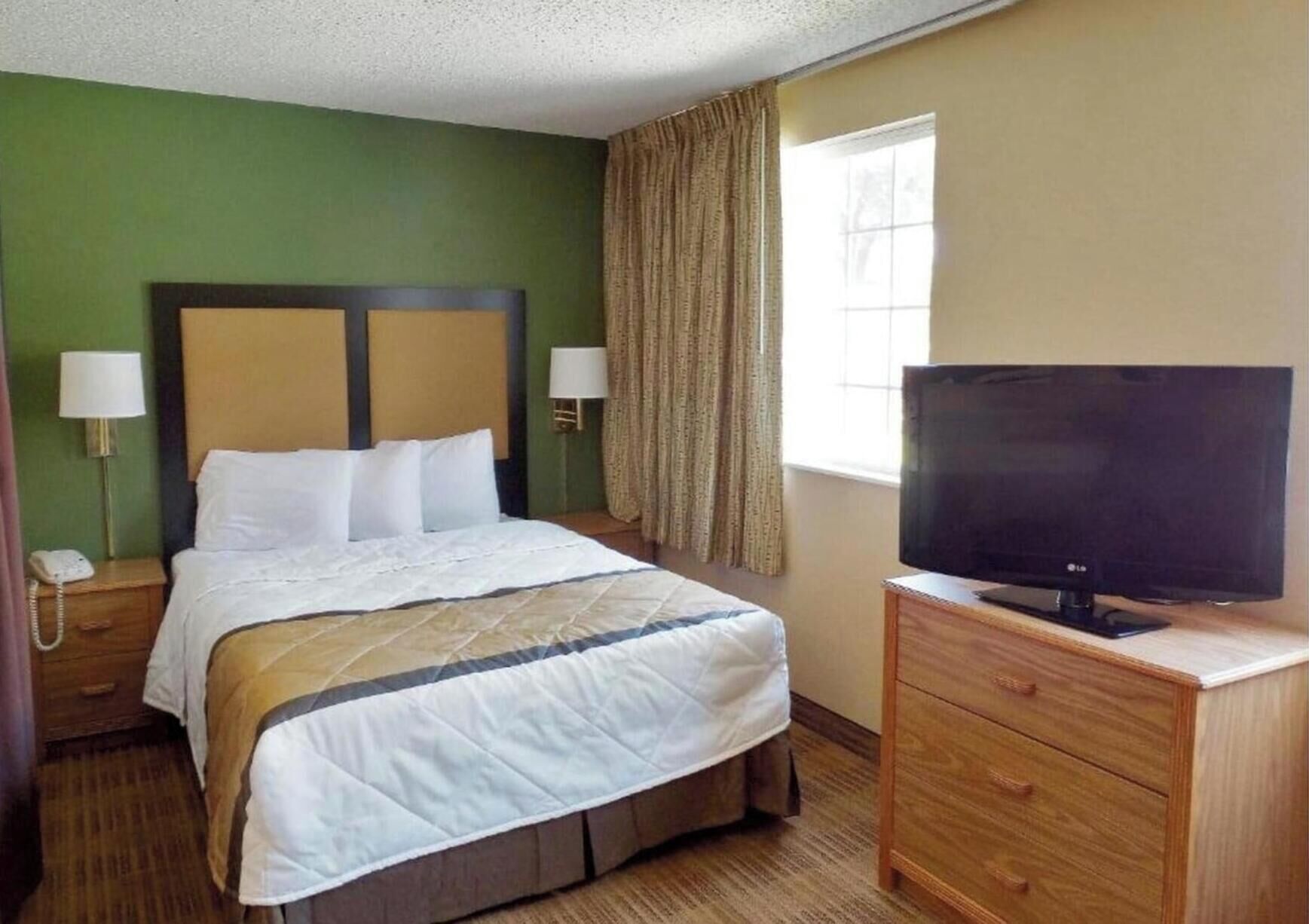 A hotel room with a bed, a TV on a wooden stand, a nightstand with a phone, and a window with curtains.