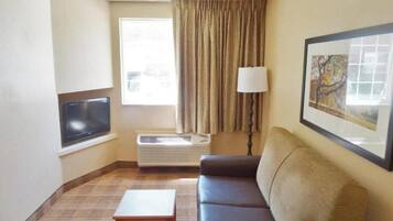 A hotel room with a sofa, a small table, a TV, a window with curtains, an air conditioner, and a lamp.