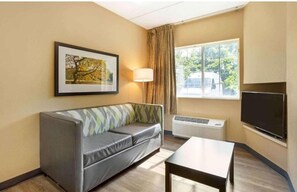Habitación de hotel con sofá, televisión de pantalla plana, lámpara y ventana con vista a árboles y un vehículo afuera.