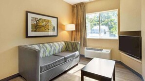 A hotel room with a sofa, a table, a TV, a window with curtains, and a framed picture on the wall. - Studio 6 Fishkill|King bed|Hearing Access|Kitchen (Fishkill)