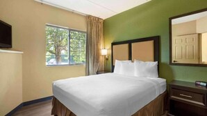 A hotel room with a bed, a window with curtains, a bedside lamp, a mirror, and a TV. - Studio 6 Fishkill|King bed|Hearing Access|Kitchen (Fishkill)