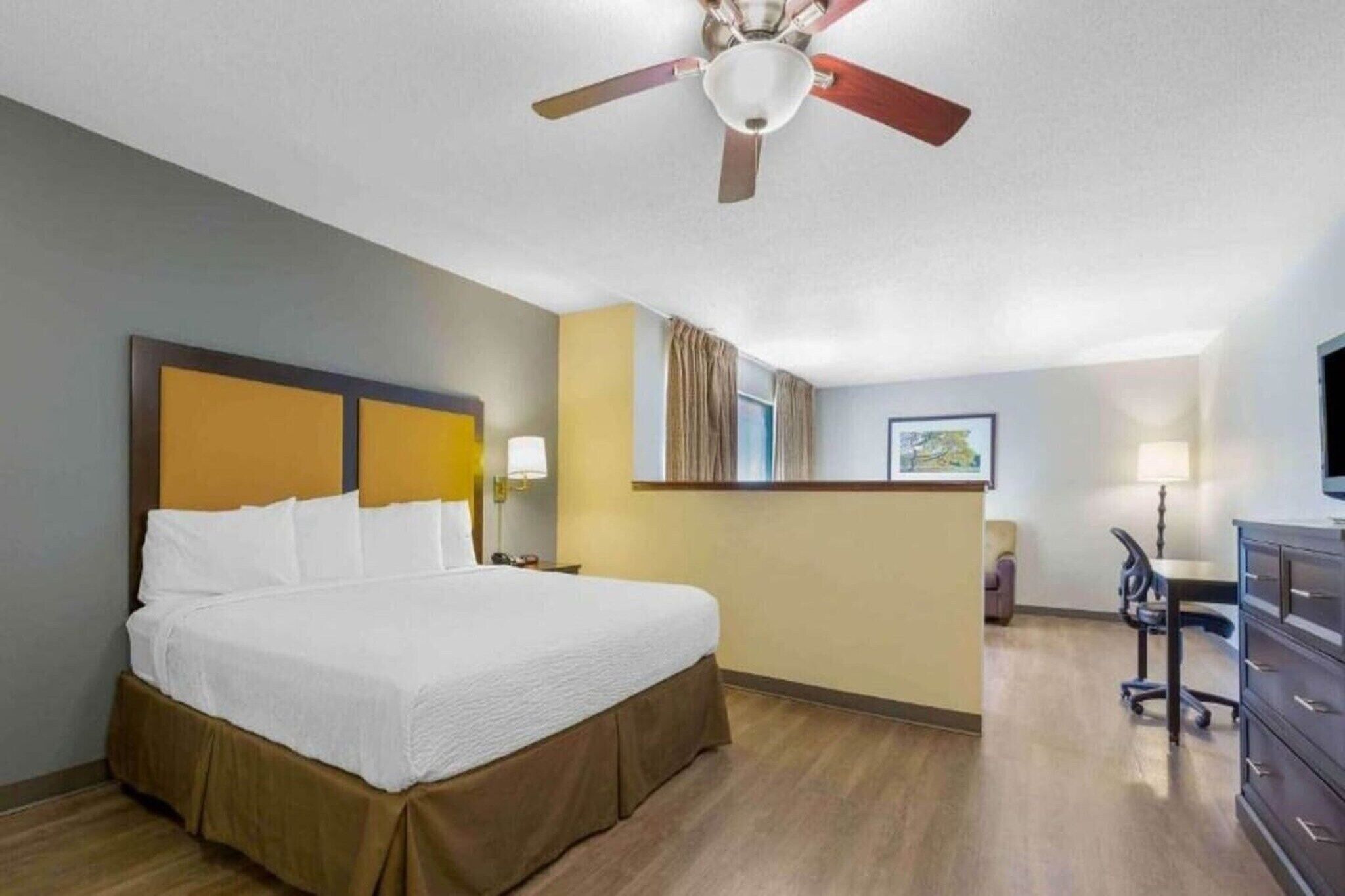 Ein Hotelzimmer mit Bett, Schreibtisch, Stuhl, Kommode, Deckenventilator und einem Fenster mit Vorhängen.